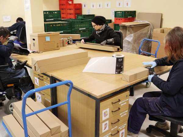 Mitarbeitende der Union Sozialer Einrichtungen verpacken Garderoben an einem Packtisch in der Werkstatt