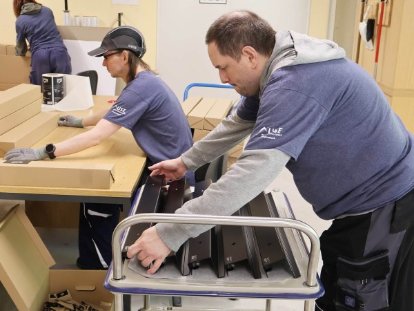 Zwei Mitarbeitende der Union Sozialer Einrichtungen nehmen schwarze Garderoben von einem Rollwagen und verpacken sie in Kartons